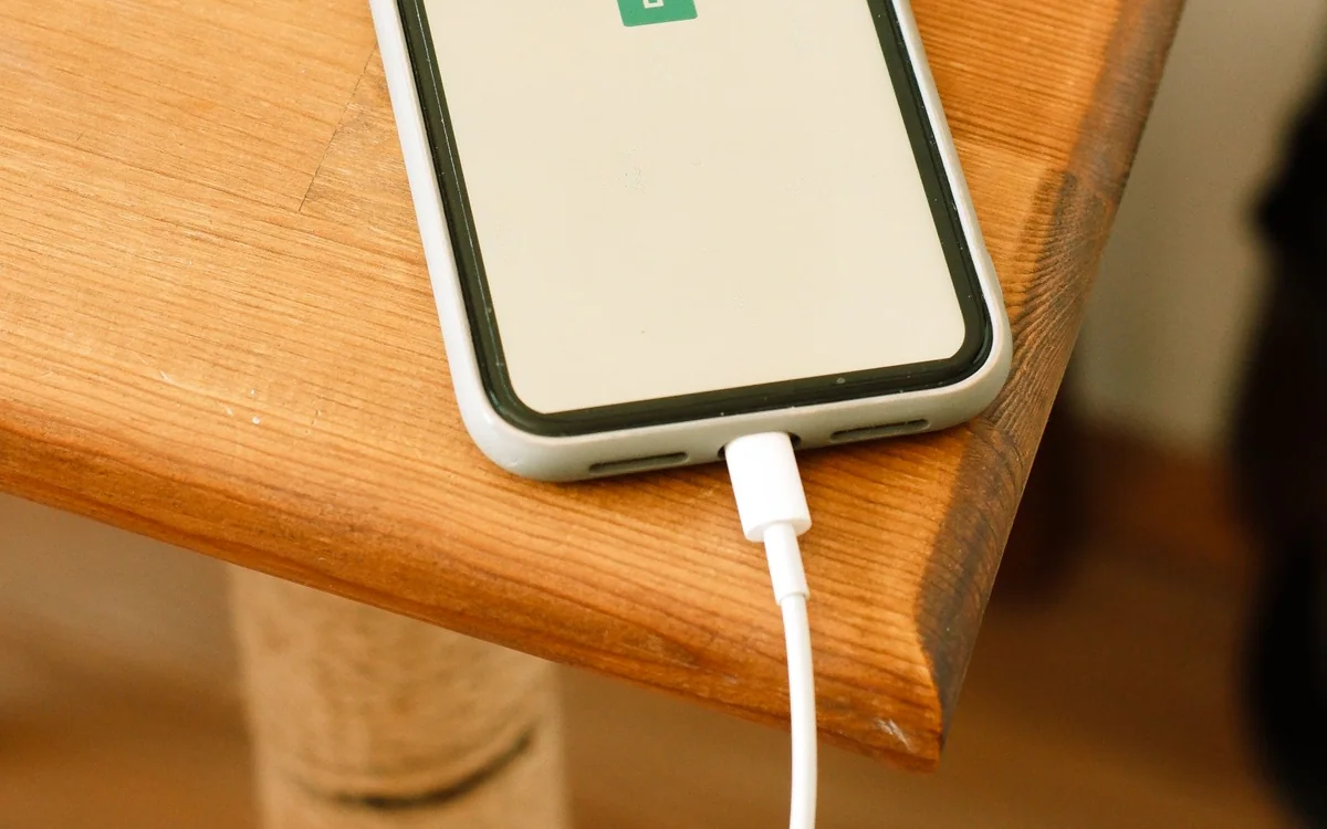 A phone charging on a bedside table with a book and a table lamp